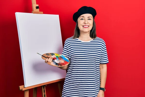 Orta yaşlı İspanyol kadın, yüzünde mutlu ve havalı bir gülümsemeyle paletle resim çiziyor. şanslı kişi. 