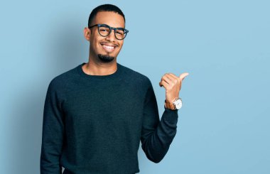Sıradan kıyafetler ve gözlük takan, gülümseyen, gülümseyen ve baş parmağıyla yüzünü gösteren Afro-Amerikan genç bir adam.. 