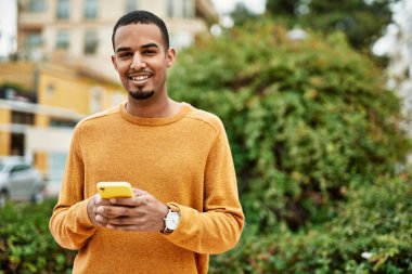 Genç Afrikalı Amerikalı adam şehirde akıllı telefon kullanarak mutlu bir şekilde gülümsüyor..