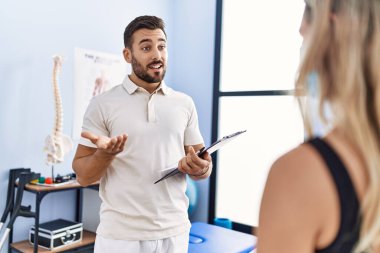 Fizyoterapist üniforması giyen genç İspanyol adam klinikteki hastayla konuşuyor.