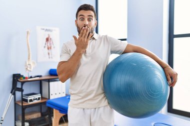Rehabilitasyon kliniğinde elinde pilates topu tutan yakışıklı bir adam ağzı elle kapatıyor, şok geçiriyor ve hata yapmaktan korkuyor. şaşırmış ifade 