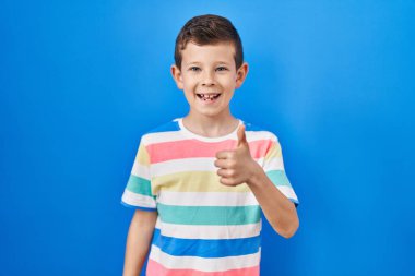 Mavi arka planda duran genç beyaz çocuk elleriyle mutlu bir jest yapıyor. Kameraya bakıp başarıyı gösteren ifade. 