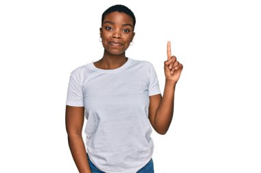 Genç Afro-Amerikalı kadın, günlük beyaz tişört giyip bir numaralı parmağını gösterirken kendine güvenen ve mutlu bir şekilde gülümsüyor.. 