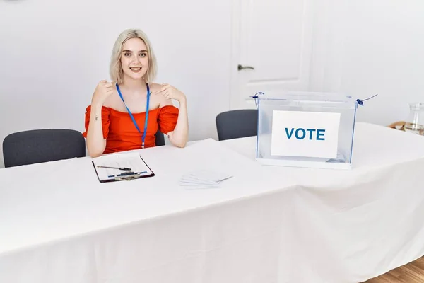 Young caucasian woman at political election sitting by ballot looking confident with smile on face, pointing oneself with fingers proud and happy.