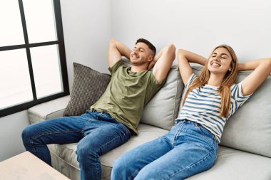 Genç İspanyol çift mutlu bir şekilde gülümsüyor. Elleri başının üstünde evdeki kanepede oturuyor..