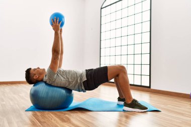 Genç İspanyol adam gülümsüyor. Spor merkezinde top kullanarak karın kasları çalıştırıyor.