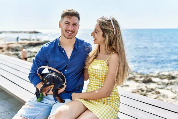 Młoda Kaukaska Para Uśmiechnięta Szczęśliwa Siedząc Ławce Psem Plaży — Zdjęcie stockowe