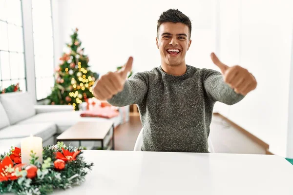 Молодий Іспаномовний Чоловік Сидить Столі Різдвяної Ялинки Схвалюючи Позитивний Жест — стокове фото