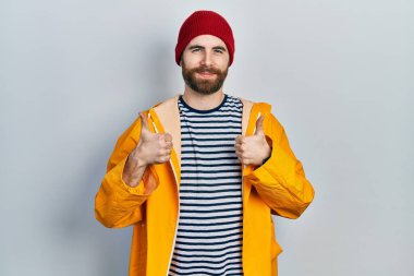 Sakallı, sarı yağmurluk giyen, elinde olumlu bir jest yapan, gülümseyen ve mutlu beyaz bir adam. Neşeli ifade ve kazanan jest. 