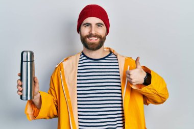 Sakallı, termosu olan beyaz bir adam mutlu ve pozitif gülümsüyor, başparmağıyla mükemmel ve onay işareti yapıyor. 