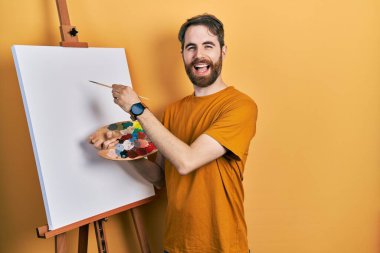 Beyaz sakallı bir adam paletle resim çiziyor. Resim sehpası ayakta dikiliyor ve kahkahalarla gülüyor.. 