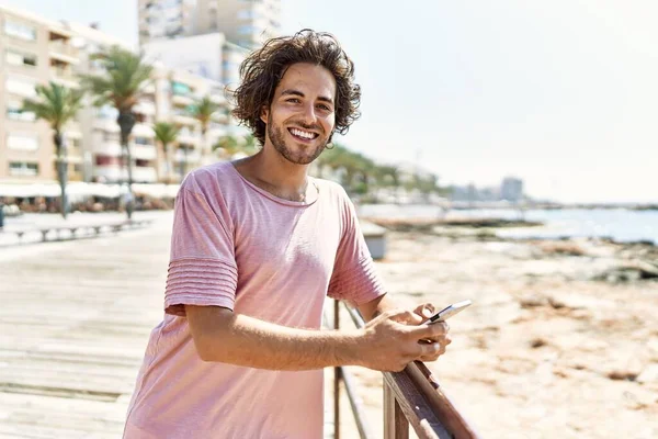 Ung Latinamerikansk Man Ler Glad Med Smartphone Stående Stranden — Stockfoto