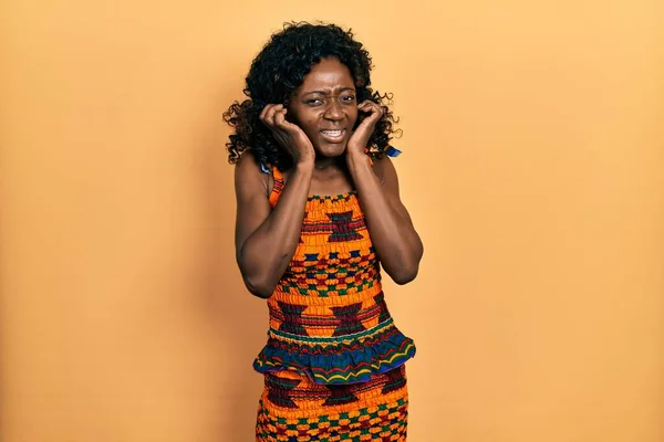 アフリカ系アメリカ人の若い女性が伝統的なアフリカの服を着て 大きな音楽のノイズに悩まされた表情で耳を覆う 聴覚障害の概念 — ストック写真