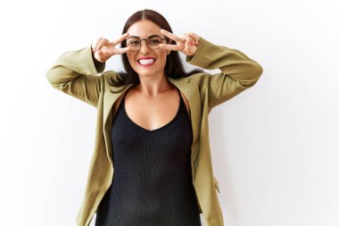 Genç esmer kadın izole edilmiş arka planda duruyor. Barış sembolü yapıyor. Yüzünde parmakları, gülümseyerek zaferi gösteriyor. 