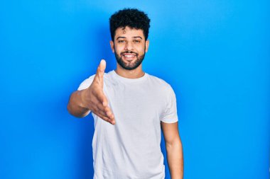 Sakallı, günlük beyaz tişörtlü genç bir Arap dostça bir selamlaşma ve selamlama sunuyor. başarılı iş. 