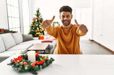 Arap genç adam Noel ağacının yanında oturmuş, el hareketleriyle olumlu jest yapmayı onaylıyor, gülümsüyor ve başarı için seviniyor. kazanan jest. 