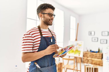 Genç Arap sanatçı gülümsüyor. Resim stüdyosunda fırça ve palet tutarken mutlu..