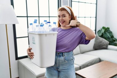 Geri dönüşüm sepetini elinde plastik şişelerle tutan genç kızıl saçlı kadın telefonla el ve parmaklarıyla telefonla konuşuyor gibi gülümsüyor. iletişim kavramları. 