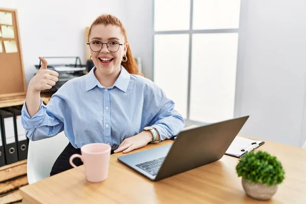 在办公室里工作的红头发年轻女人用电脑笔记本电脑做着快乐的大拇指举手表决 赞成的表情看着相机显示的成功 — 图库照片