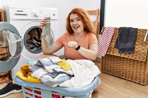Kirli çamaşırlarını çamaşır makinesine koyan genç, kızıl saçlı bir kadın arka tarafı işaret ediyor. Elleri havada, kendinden emin bir şekilde. 