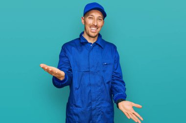 Sakallı kel adam, inşaatçı tulumu giyiyor, neşeli gülümsüyor, dostça karşılanıyor, olumlu ve kendinden emin selamlar. 