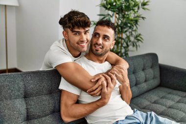 İki İspanyol erkek, kendilerine güvenerek gülümsüyor ve evde birbirlerine sarılıyorlar.