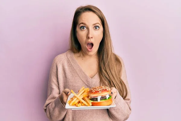 若いですブロンド女性食べるおいしいです古典的なハンバーガーとともにフライドポテト恐れと衝撃で驚きと驚きの表情 恐怖と興奮した顔 — ストック写真