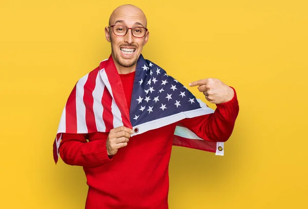 Bald man with beard wrapped around united states flag smiling happy pointing with hand and finger