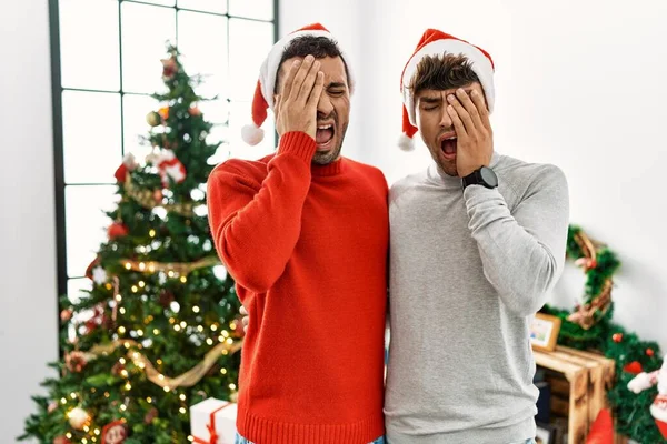 若いゲイのカップルは 手で半分の顔 目と口をカバー疲れてあくび帽子をかぶってクリスマスツリーで立っている 痛みで顔が痛い — ストック写真