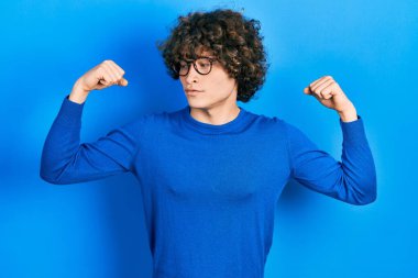 Sıradan kıyafetler ve gözlük takan yakışıklı genç adam gururla gülümseyen kol kaslarını gösteriyor. fitness konsepti. 