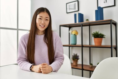 Genç Çinli kız gündelik giysiler içinde evde masada oturmuş mutlu ve pozitif görünüyor. Kendine güvenen bir gülümsemeyle dişlerini gösteriyor. 