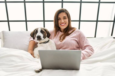 Yatak odasında dizüstü bilgisayar kullanan, köpeğini kucaklayan genç bir kadın.