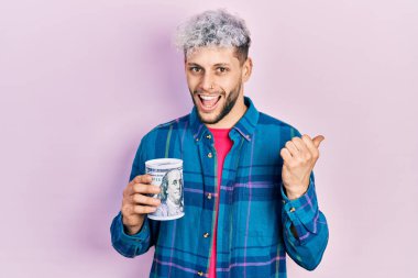 Modern boyalı saçları olan genç İspanyol bir adam domuz kumbarası tutuyor. Parmağını kenarda tutuyor ve ağzı açık gülümsüyor. 