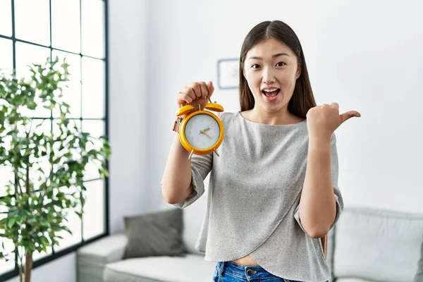 Joven Asiática Mujer Sosteniendo Alarma Reloj Apuntando Pulgar Hasta Lado — Foto de Stock