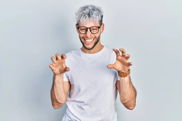 Young hispanic man with modern dyed hair wearing white t shirt and glasses smiling funny doing claw gesture as cat, aggressive and sexy expression