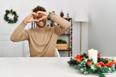 Masada sakallı genç yakışıklı adam Noel süslemeleri ile oturuyor. El ve parmaklarıyla kalp şekli yapıyor. Tabelanın içinden bakıp gülümsüyor. 