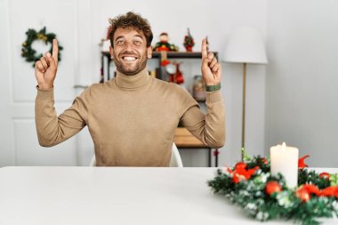Yılbaşı süslemeleriyle masada oturan sakallı yakışıklı genç adam şaşırmış, şaşırmış ve parmaklarıyla işaret ediyor ve kollarını kaldırıyordu.. 