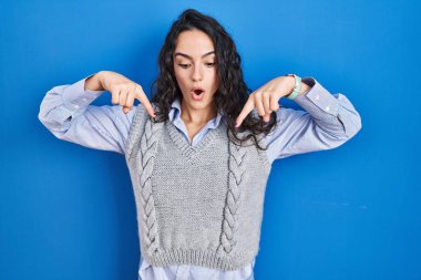 Genç esmer kadın mavi arka planda dikiliyor parmaklarını göstererek reklamını yapıyor, şaşırmış yüzünü ve ağzını açıyor. 