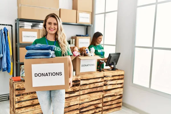 Two volunteers woman working at charity center. Girl smiling happy and holding box with clothes to donate.