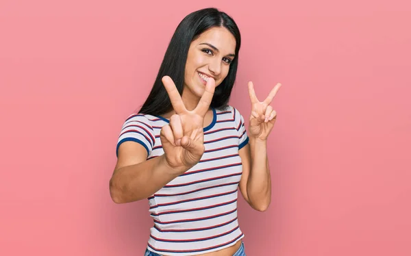 若いヒスパニック系の女の子は カジュアルな縞模様のTシャツを着てカメラを見て笑みを浮かべて勝利のサインを行う指を示す — ストック写真