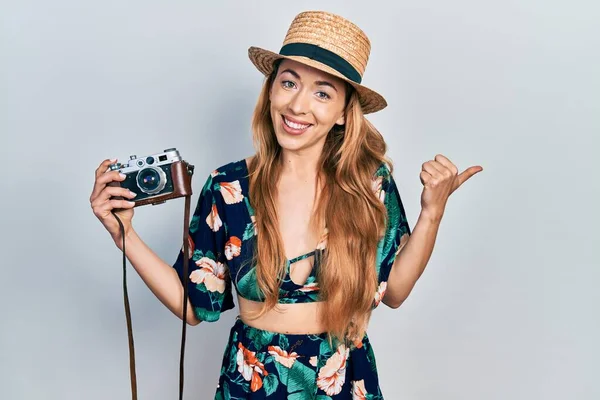 Young caucasian woman holding vintage camera pointing thumb up to the side smiling happy with open mouth