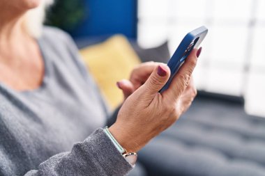 Orta yaşlı sarışın kadın akıllı telefon kullanıyor. Evdeki koltukta oturuyor.