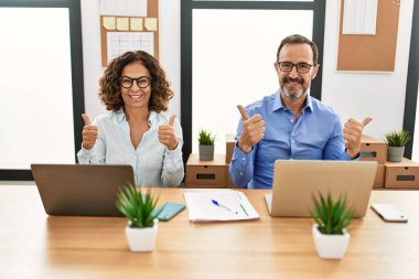 Orta yaşlı İspanyol kadın ve erkek ofis başarı tabelasında dizüstü bilgisayarlarıyla oturup el hareketleriyle olumlu jest yapıyorlar, gülümsüyorlar ve mutlular. Neşeli ifade ve kazanan jest. 
