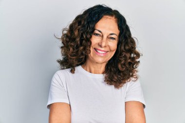 Orta yaşlı İspanyol kadın, günlük beyaz tişört giyip kollarını kavuşturup kameraya bakarken gülümsüyor. pozitif kişi. 