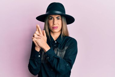 Kovboy kız tarzı giyinmiş genç İspanyol bir kadın elleri kuşkucu ve gergin bir şekilde tabanca jesti yapıyor, sorun yüzünden suratını asıyor. negatif kişi. 