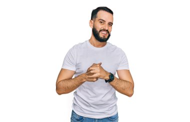 Sakallı, sıradan beyaz tişörtlü, elleri çapraz parmaklı rahat ve neşeli gülümseyen genç bir adam. Başarı ve iyimserlik 