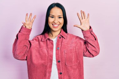 Güzel İspanyol kadın burun piercingli, günlük pembe ceket giyen, 8 numaralı parmaklarıyla işaret eden, kendinden emin ve mutlu bir şekilde gülümseyen.. 