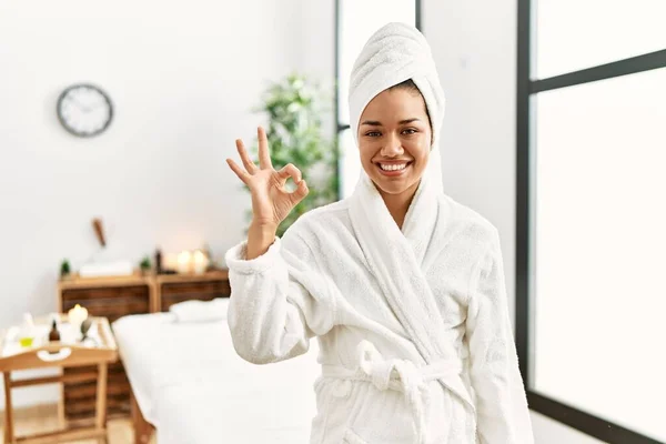 Young brunette woman wearing towel and bathrobe standing at beauty center smiling positive doing ok sign with hand and fingers. successful expression.