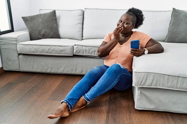 Young african woman using smartphone sitting on the floor at home laughing and embarrassed giggle covering mouth with hands, gossip and scandal concept