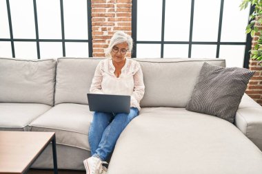 Orta yaşlı bir kadın evdeki koltukta dizüstü bilgisayar kullanıyor.